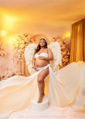 Pregnant woman with angel wings and halo poses among white flowers in ethereal maternity portrait.