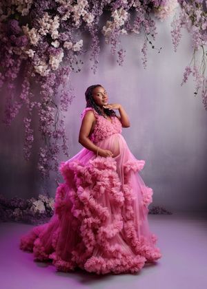 Pregnant woman in pink ruffled tulle gown poses under hanging flowers in maternity portrait.