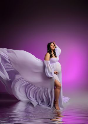 Pregnant woman in flowing lavender gown with billowing fabric stands in water for dreamy maternity portrait.