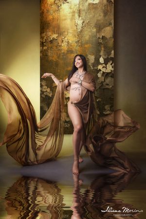 Elegant pregnant woman draped in flowing golden fabric poses in artistic maternity shoot with reflective floor.