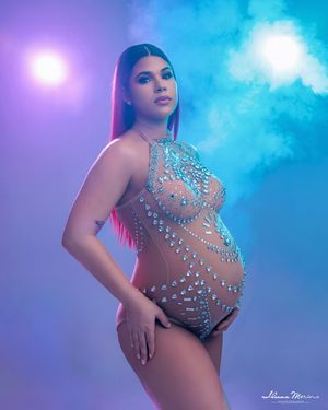 Pregnant woman wearing a crystal-embellished nude bodysuit poses in dramatic blue and purple smoke backdrop.