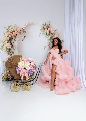 Pregnant woman in pink ruffled gown poses beside a floral baby carriage in a romantic maternity photo.