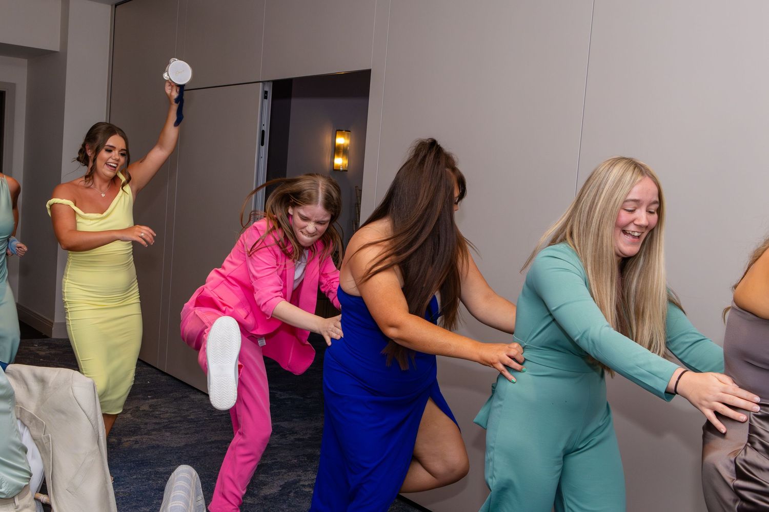 A group of people dance in a line formation wearing colorful outfits in an indoor space.