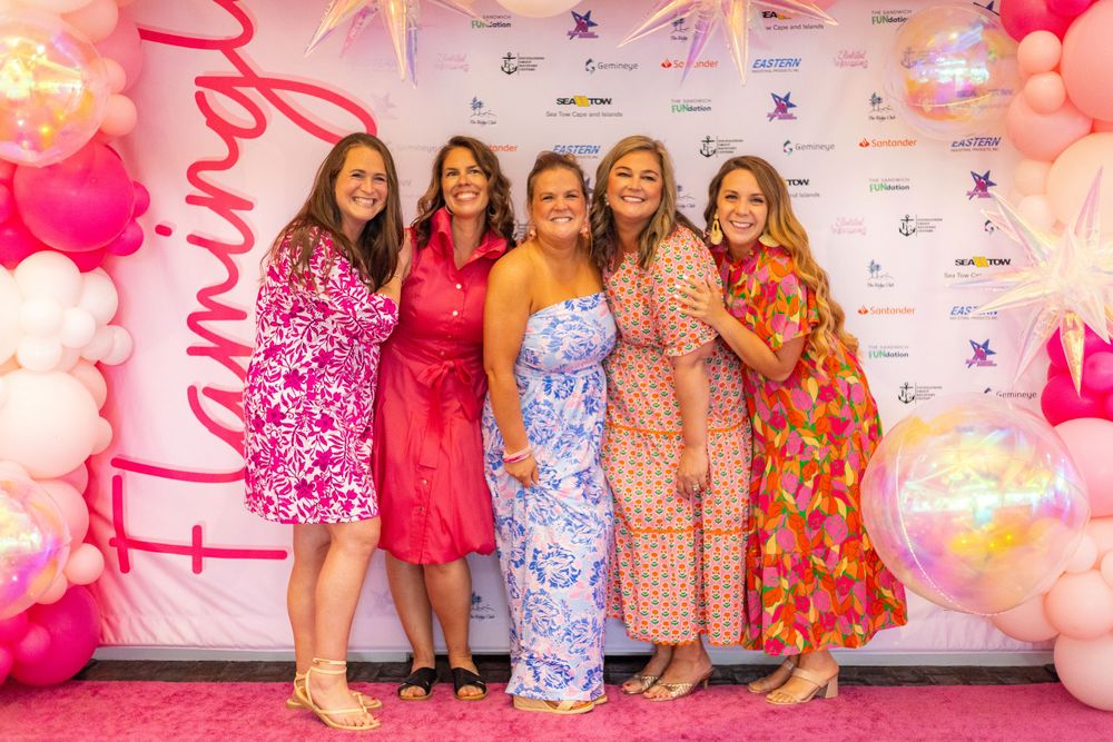 Group poses in front of pink step-and-repeat backdrop at social event with decorative balloons and lighting.