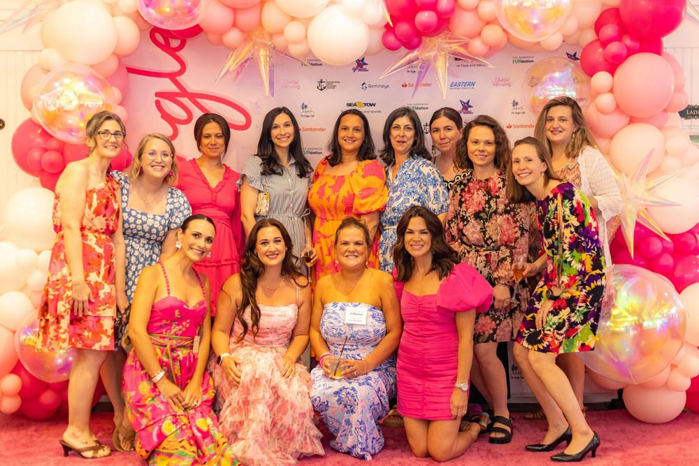A group celebrates at a pink and white balloon-filled party with decorative floral arrangements and festive attire.