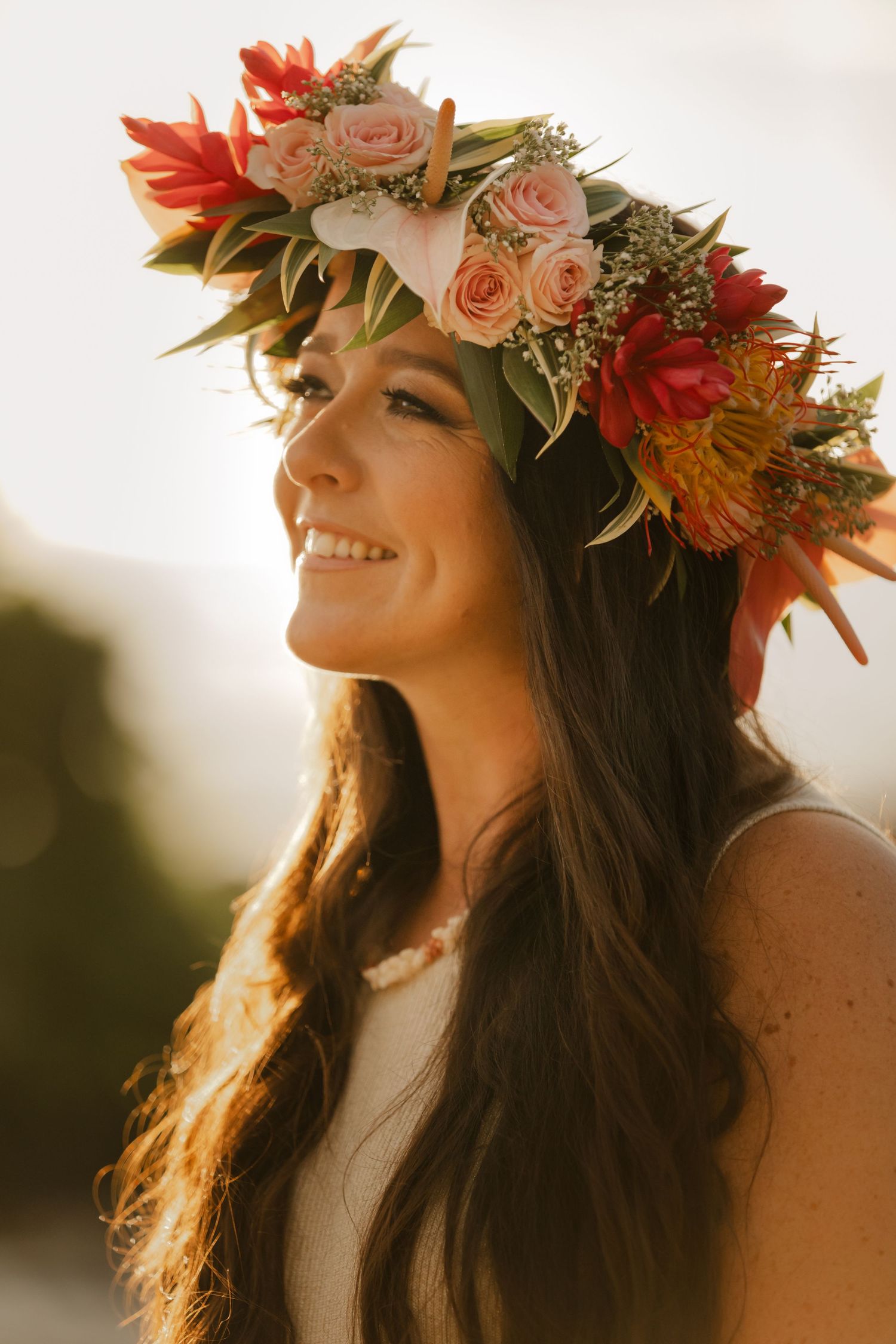 A woman wearing a floral crown of roses and tropical flowers smiles in golden sunlight during a sunset photoshoot.