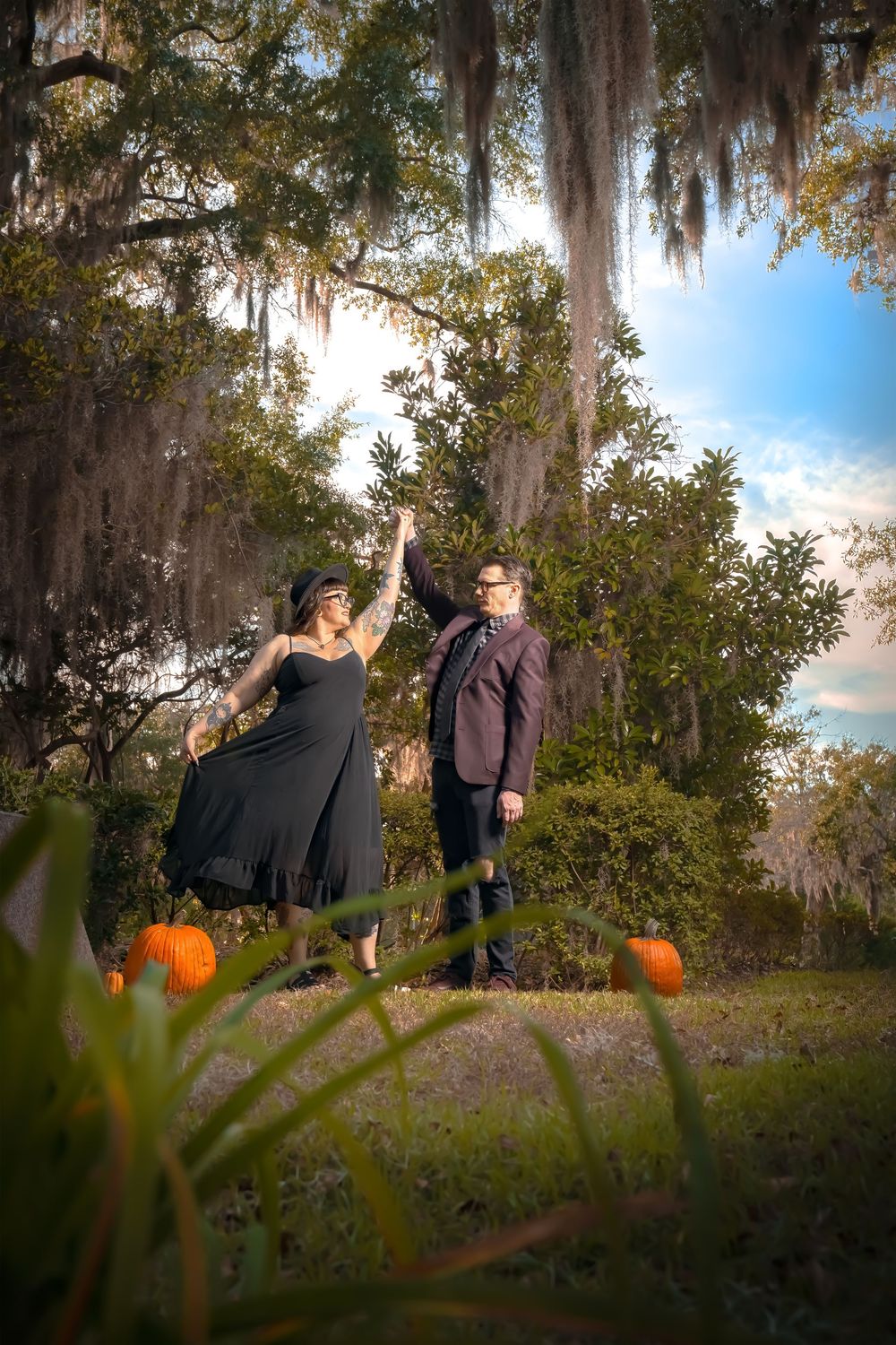 People pose near large orange pumpkins in a scenic outdoor setting with Spanish moss hanging from trees.