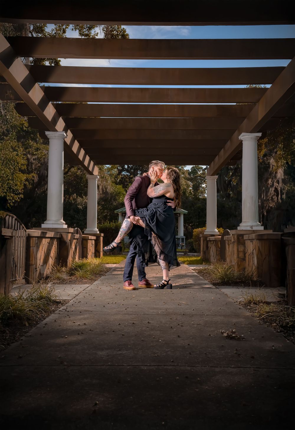 A couple shares an intimate moment under a columned pergola in an outdoor garden setting at sunset.