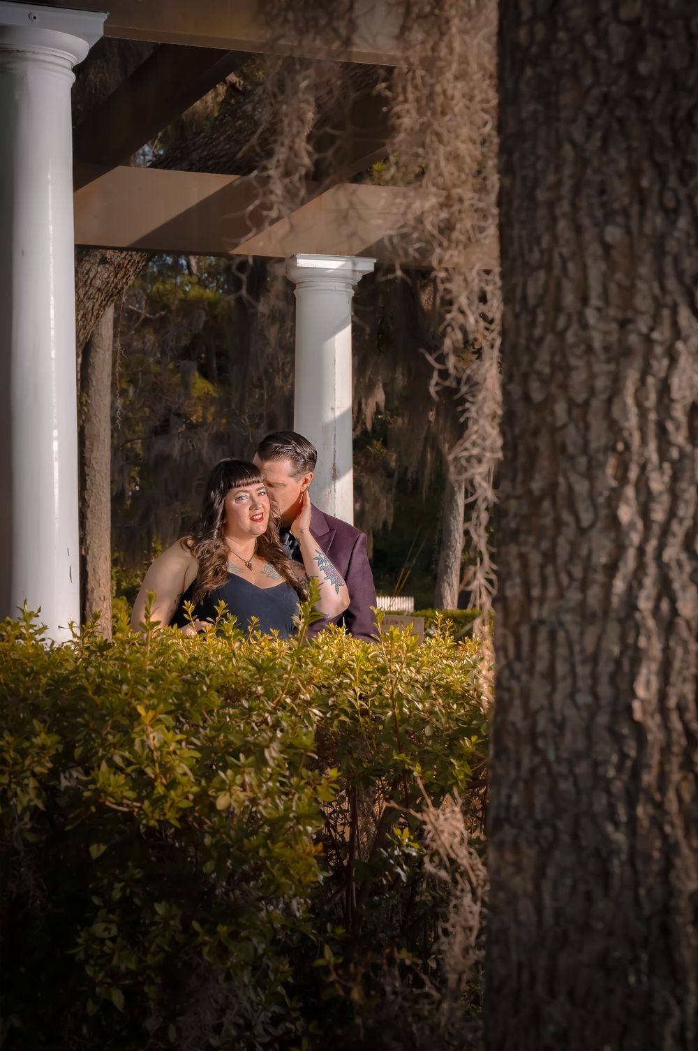 A couple shares an intimate moment at sunset behind bushes with white columns visible in the background.