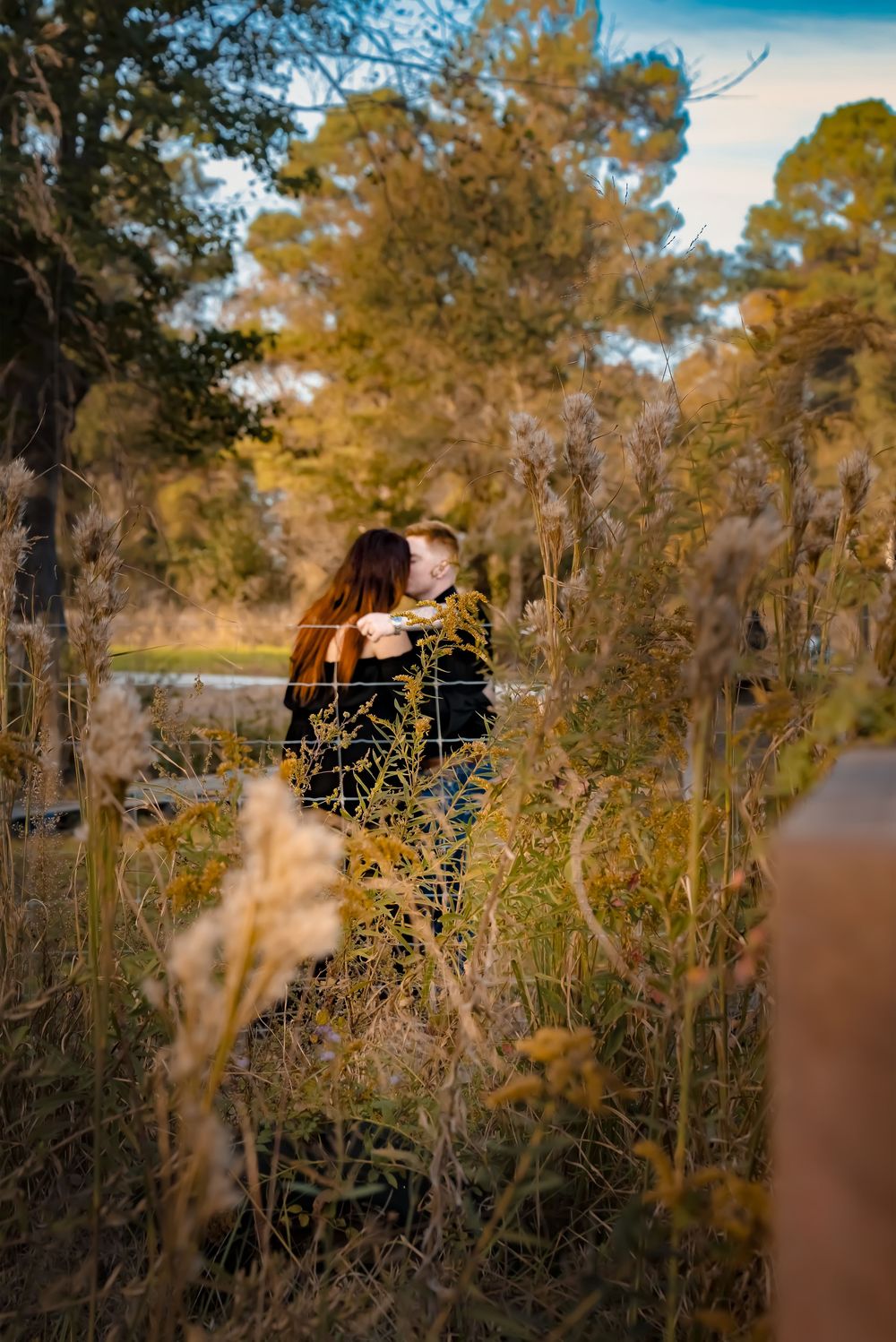 A romantic moment captured through tall grass and wildflowers at sunset in a natural outdoor setting.