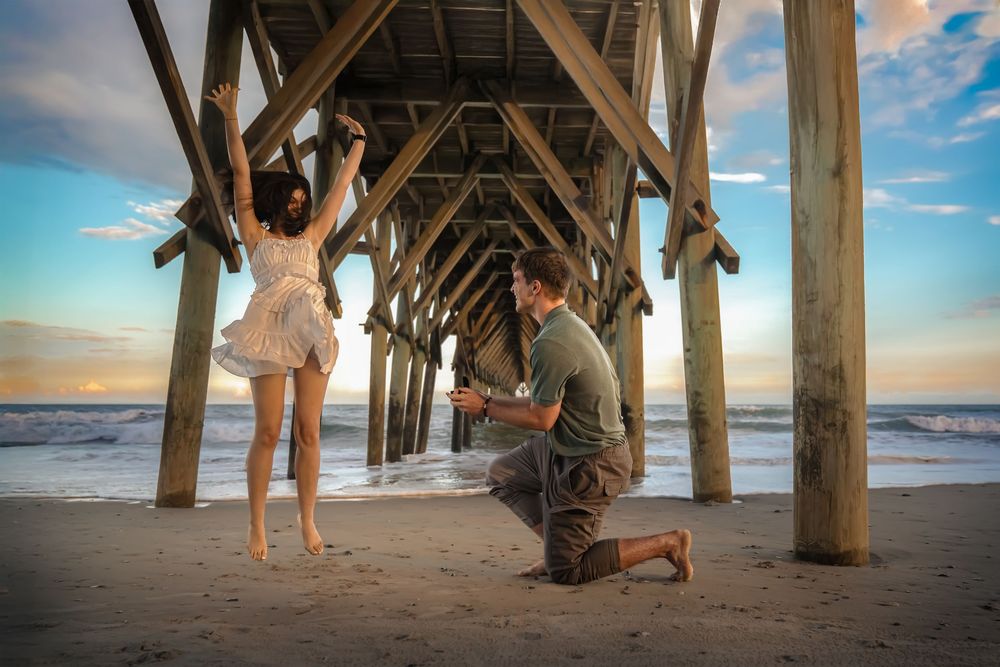 A romantic proposal moment under a wooden pier at sunset with ocean waves and beach in the background.