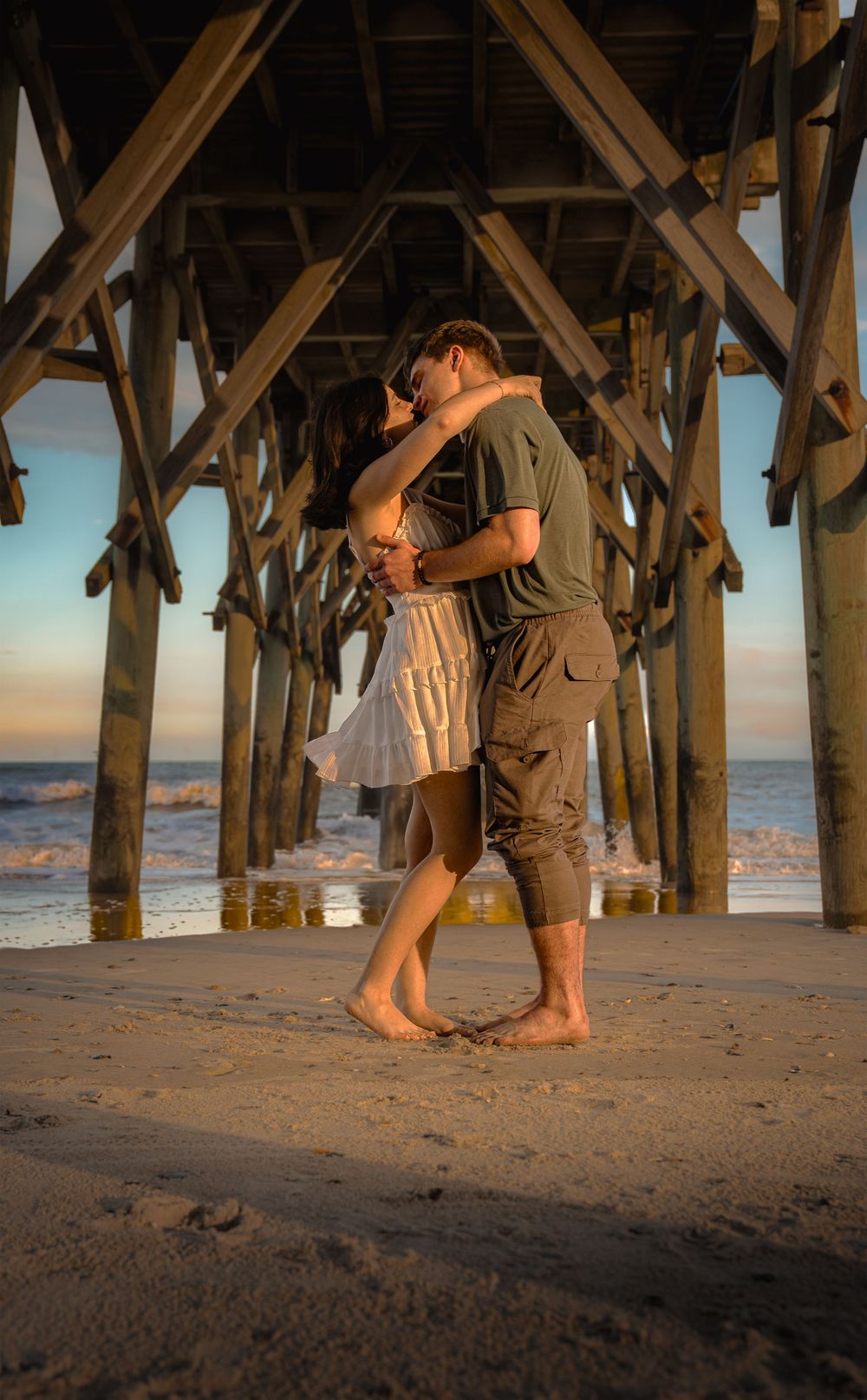 A romantic couple embraces under a wooden pier at sunset while standing barefoot in the sand on the beach.