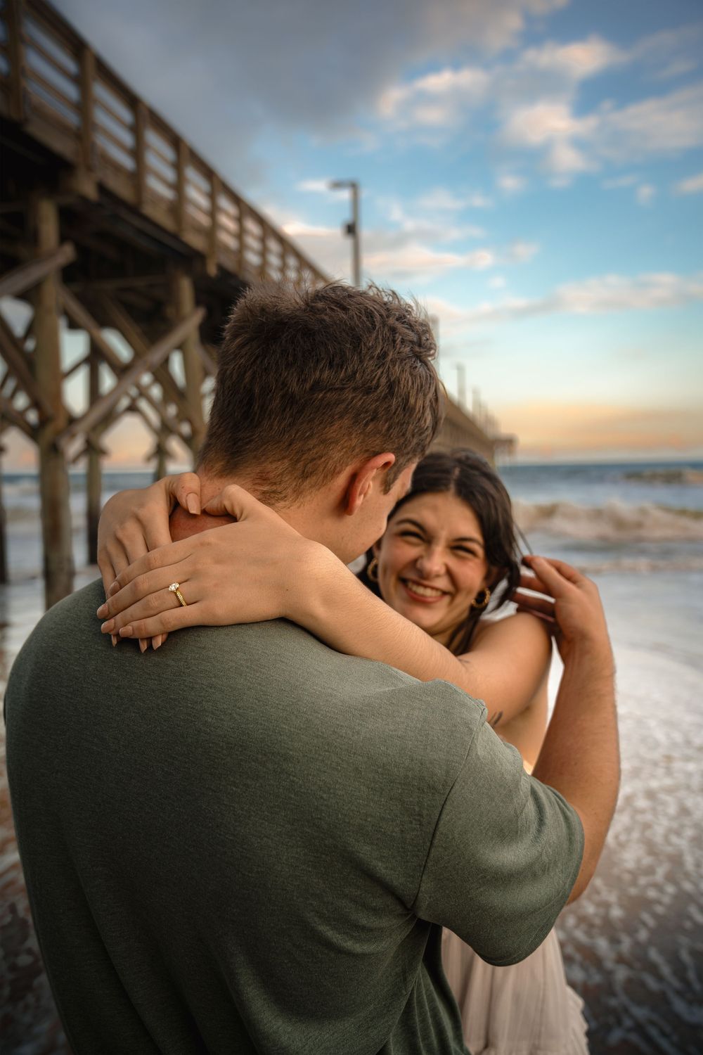 A couple embraces on a beach pier during golden hour as waves crash on the shore.