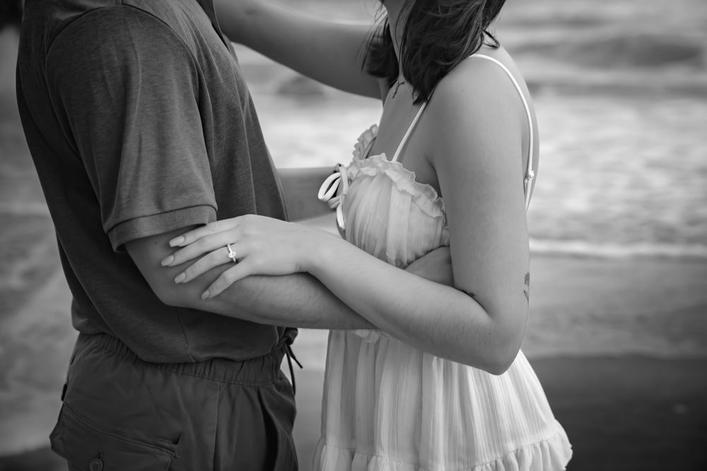 Black and white romantic beach photoshoot showing couple embracing by the ocean at sunset.