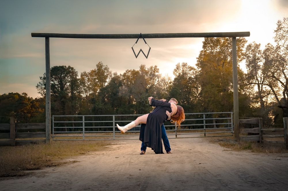 A romantic sunset dance scene at a rustic ranch entrance with a wooden archway and metal fence.
