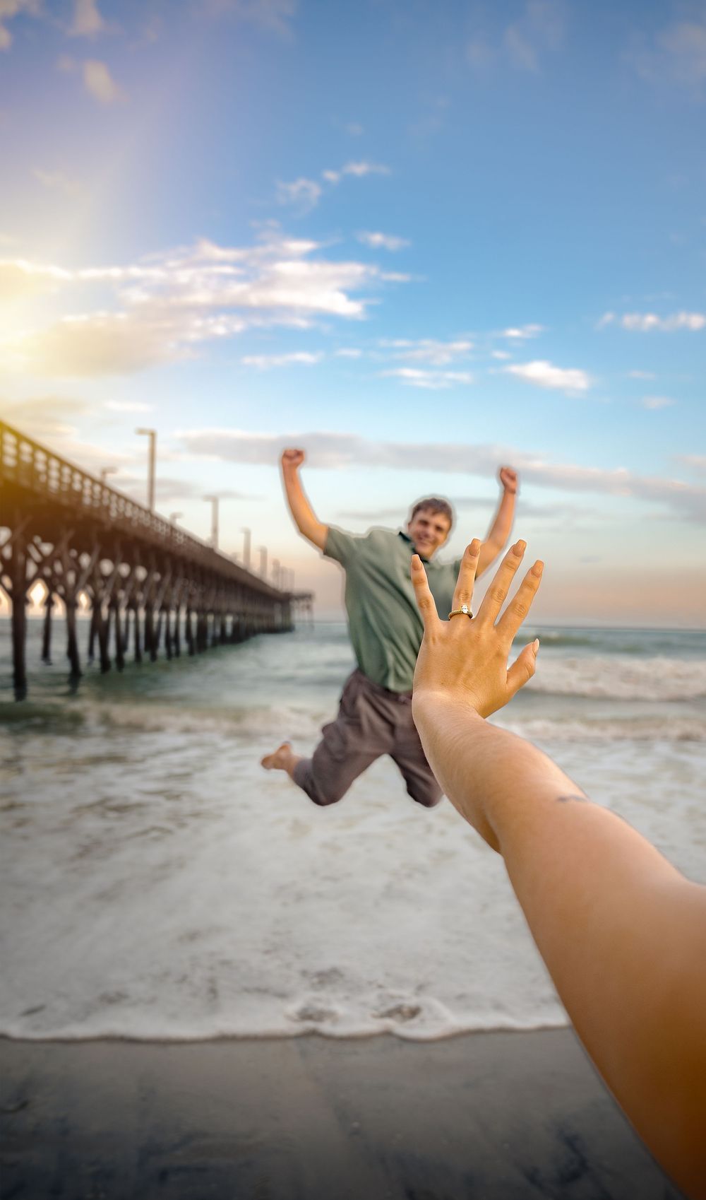 Creative forced perspective photo shows someone jumping joyfully by a beach pier while a hand reaches toward them.