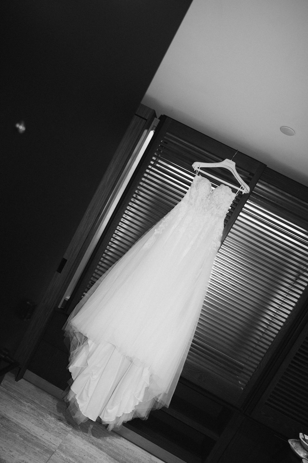 A white wedding dress hanging on display against window blinds in black and white photography.