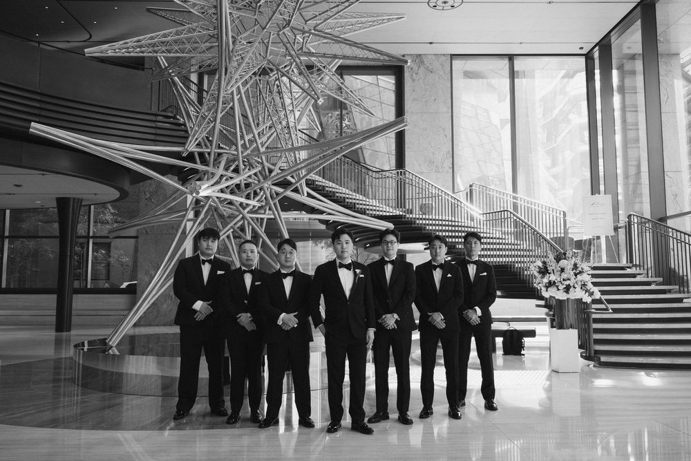Groomsmen in black tuxedos pose together in front of modern art installation.