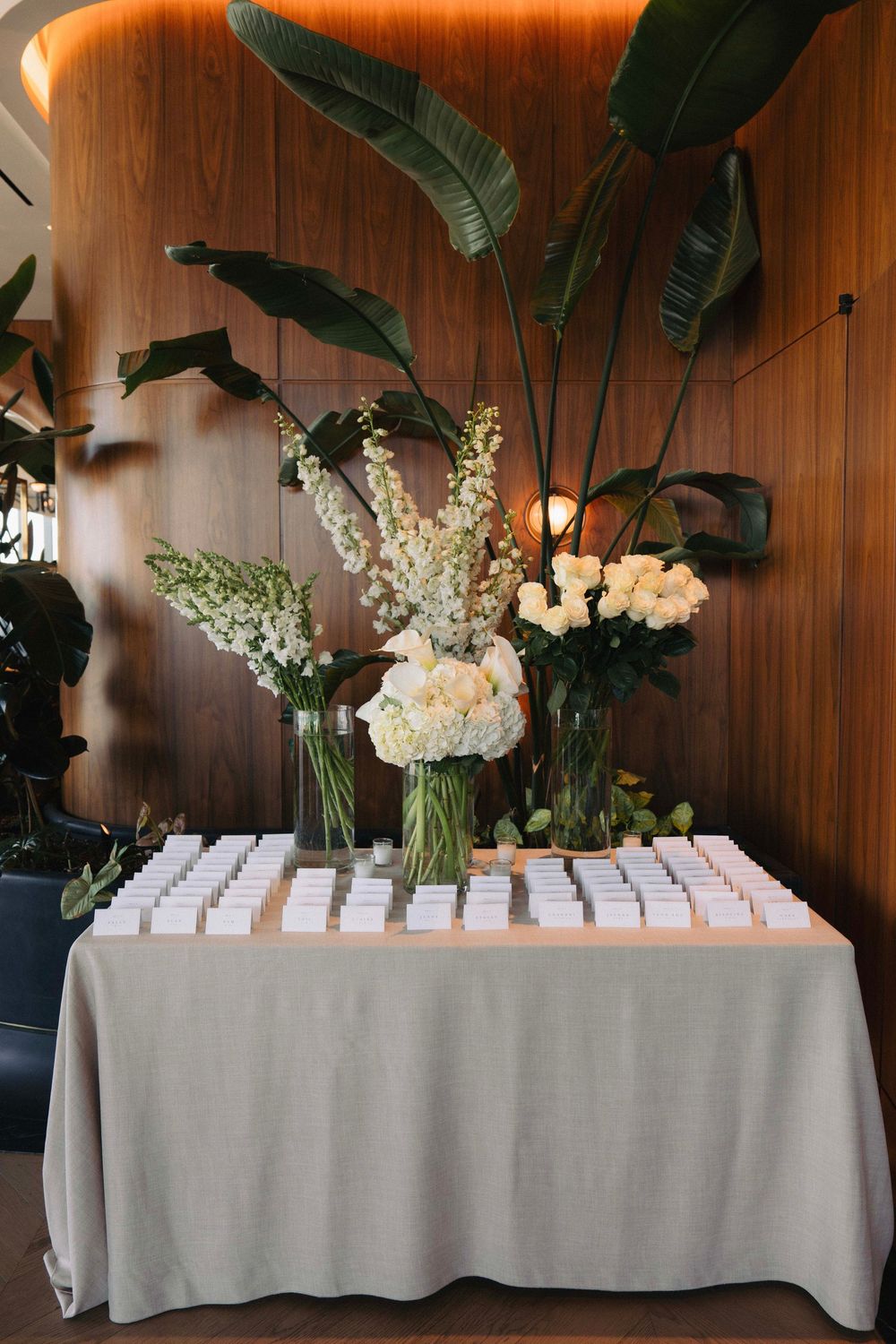 Elegant wedding seating chart display with white flowers and tropical leaves against wood paneled wall.