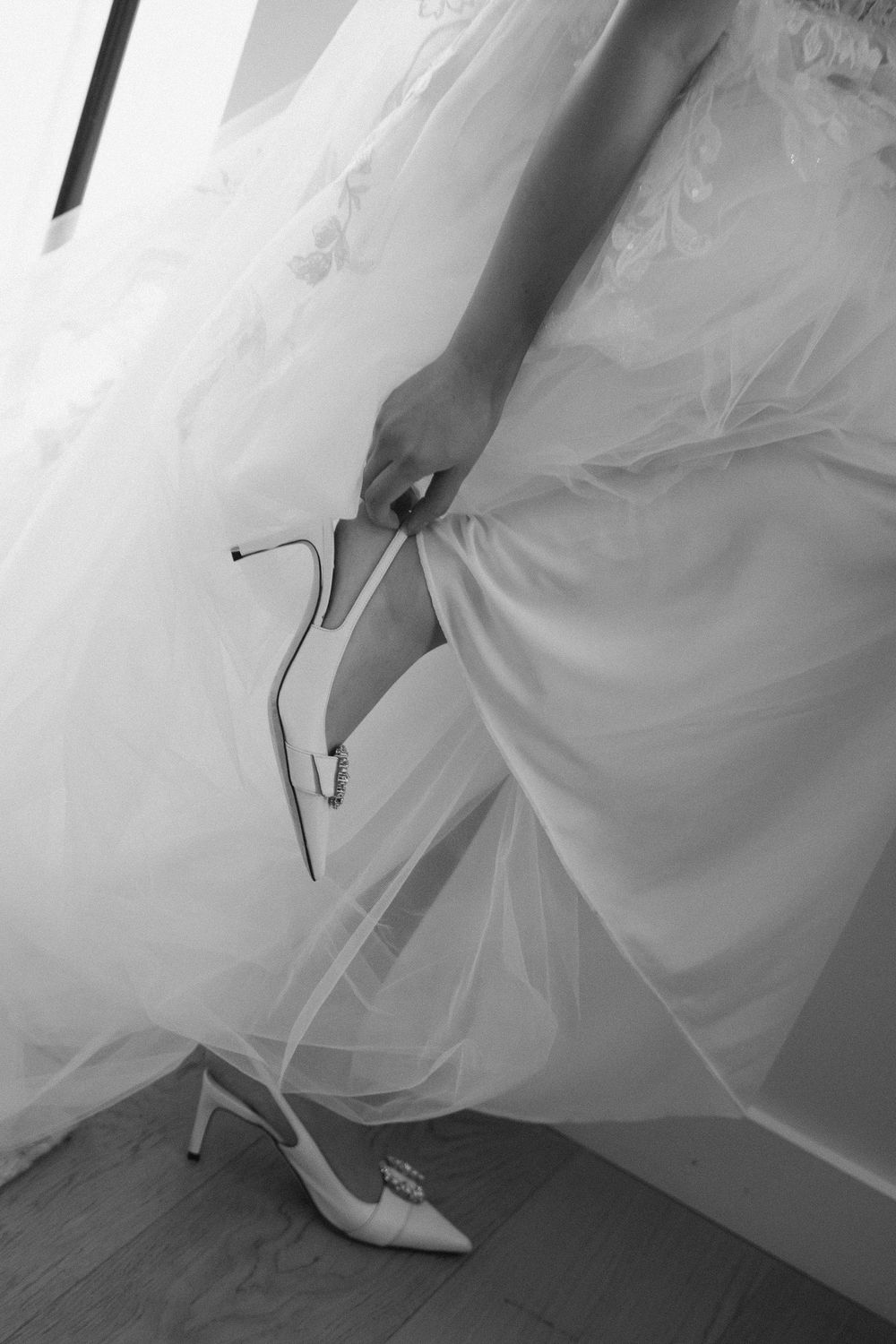 Close-up black and white photo of a bride putting on white heels with her wedding dress.
