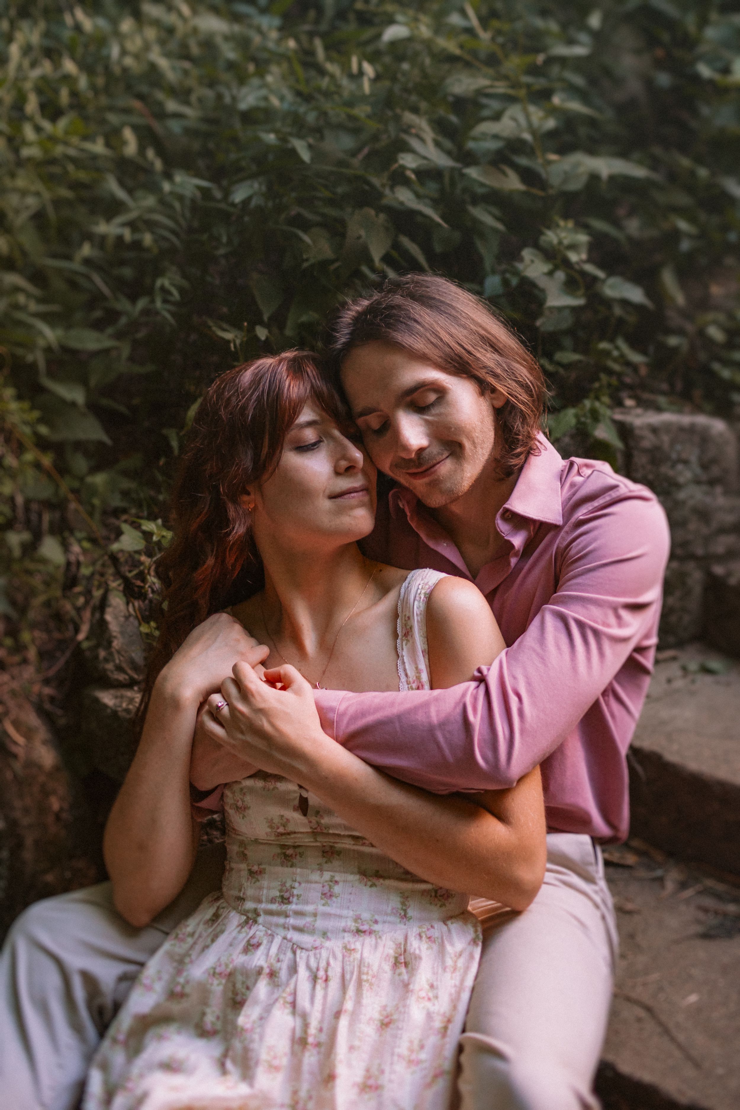 Couple in pink and white outfits sharing intimate moment among greenery.