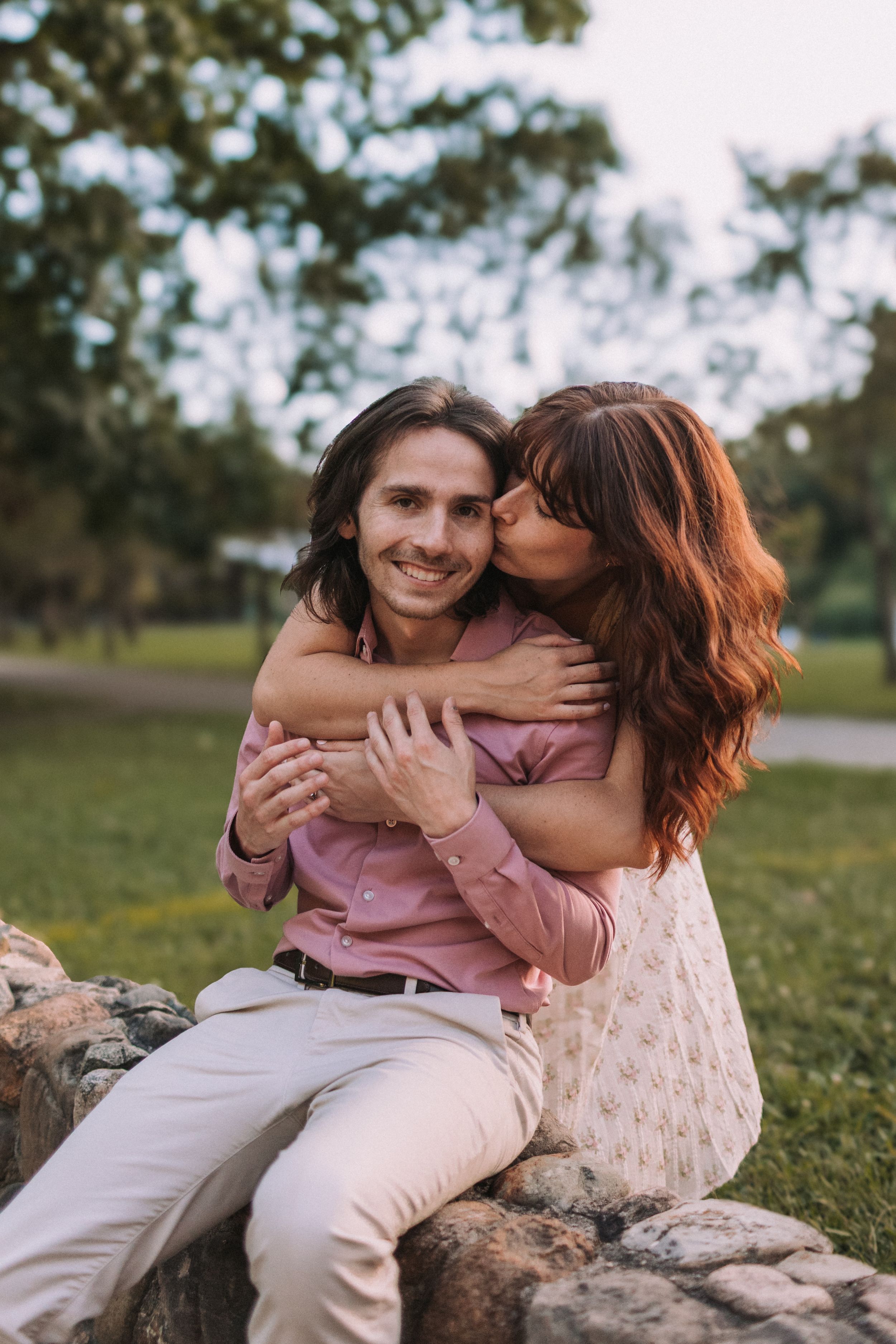 A couple sits together on rocks in a grassy field during golden hour, sharing an intimate moment in nature.