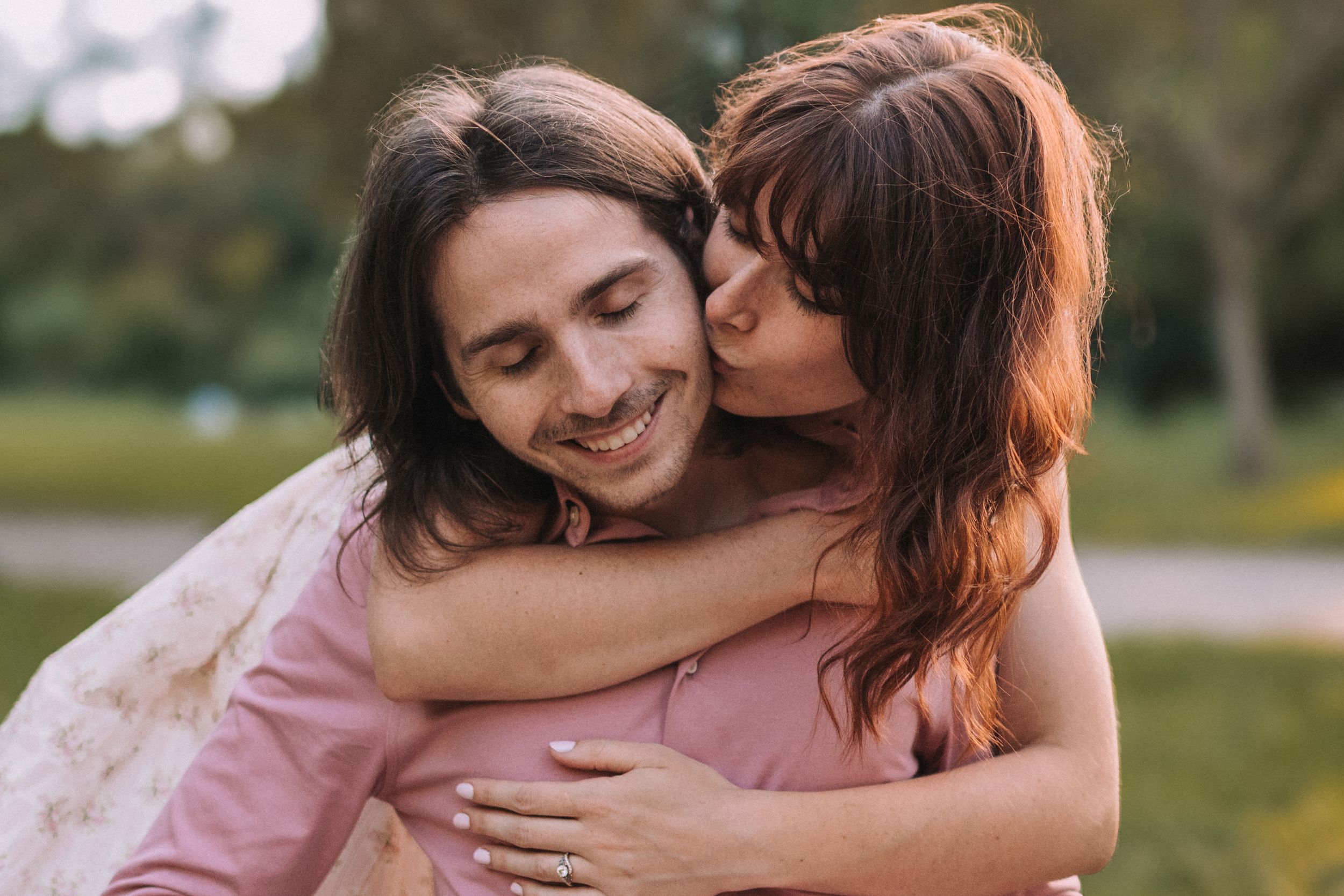 Two people share a tender embrace outdoors in a natural setting with warm sunlight filtering through the scene.