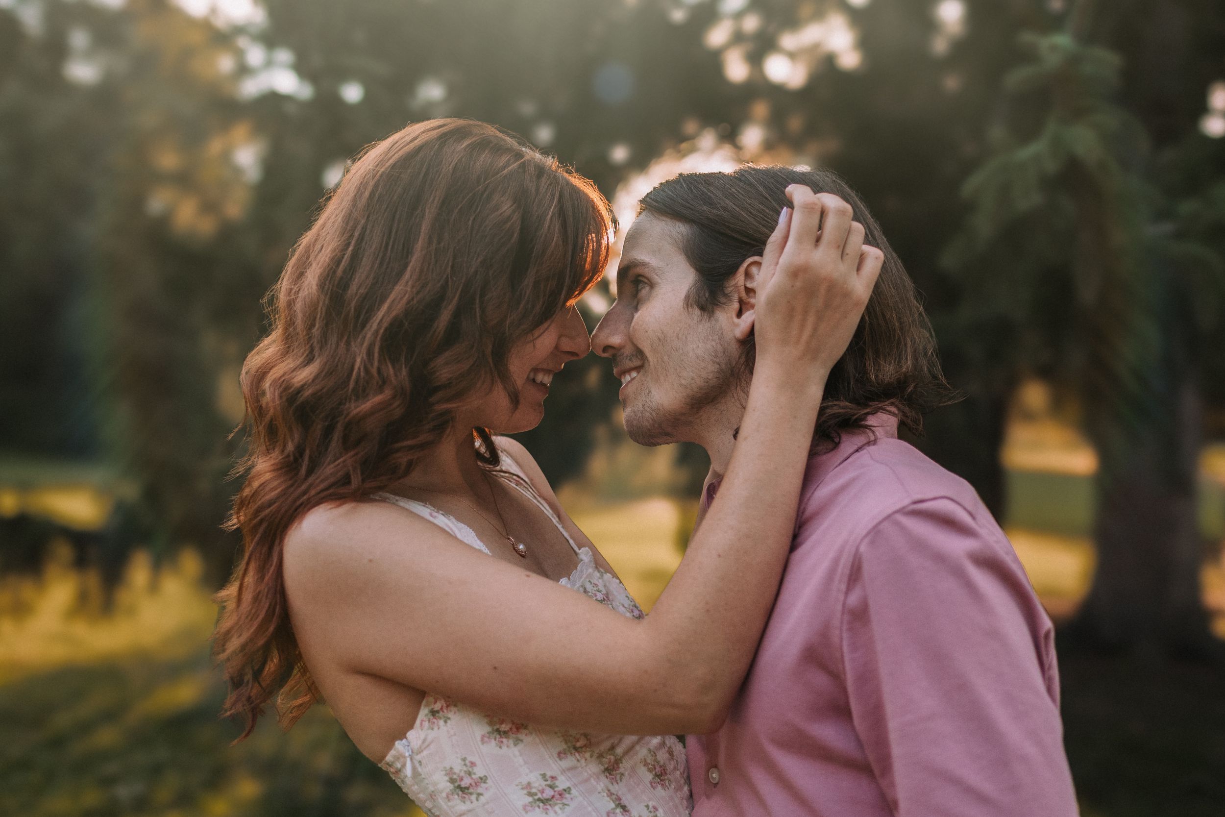 Couple in pink and white outfits embrace and kiss during sunset in a romantic outdoor setting.