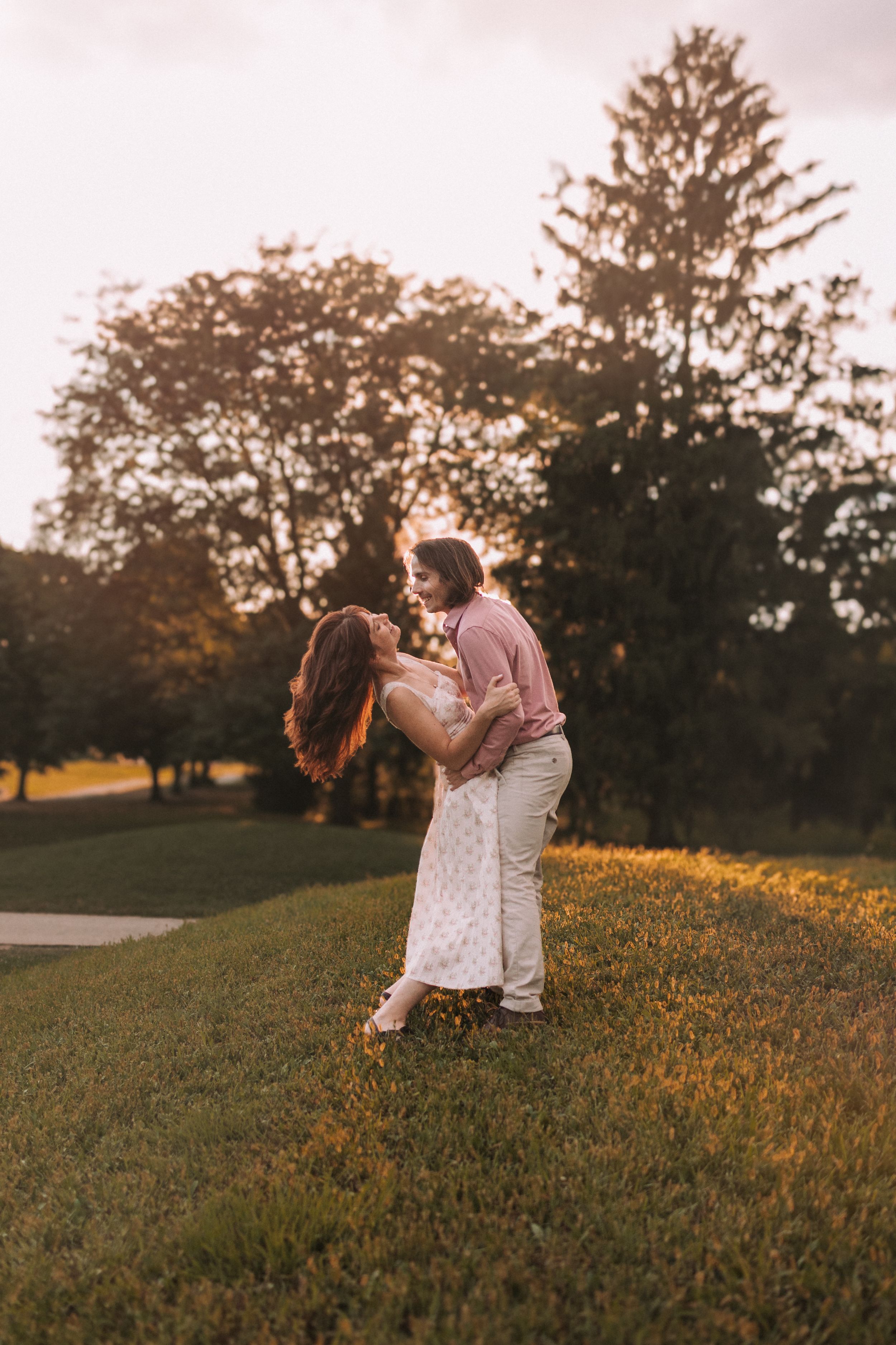 A couple embraces in a romantic moment on a grassy hillside during golden hour sunset.