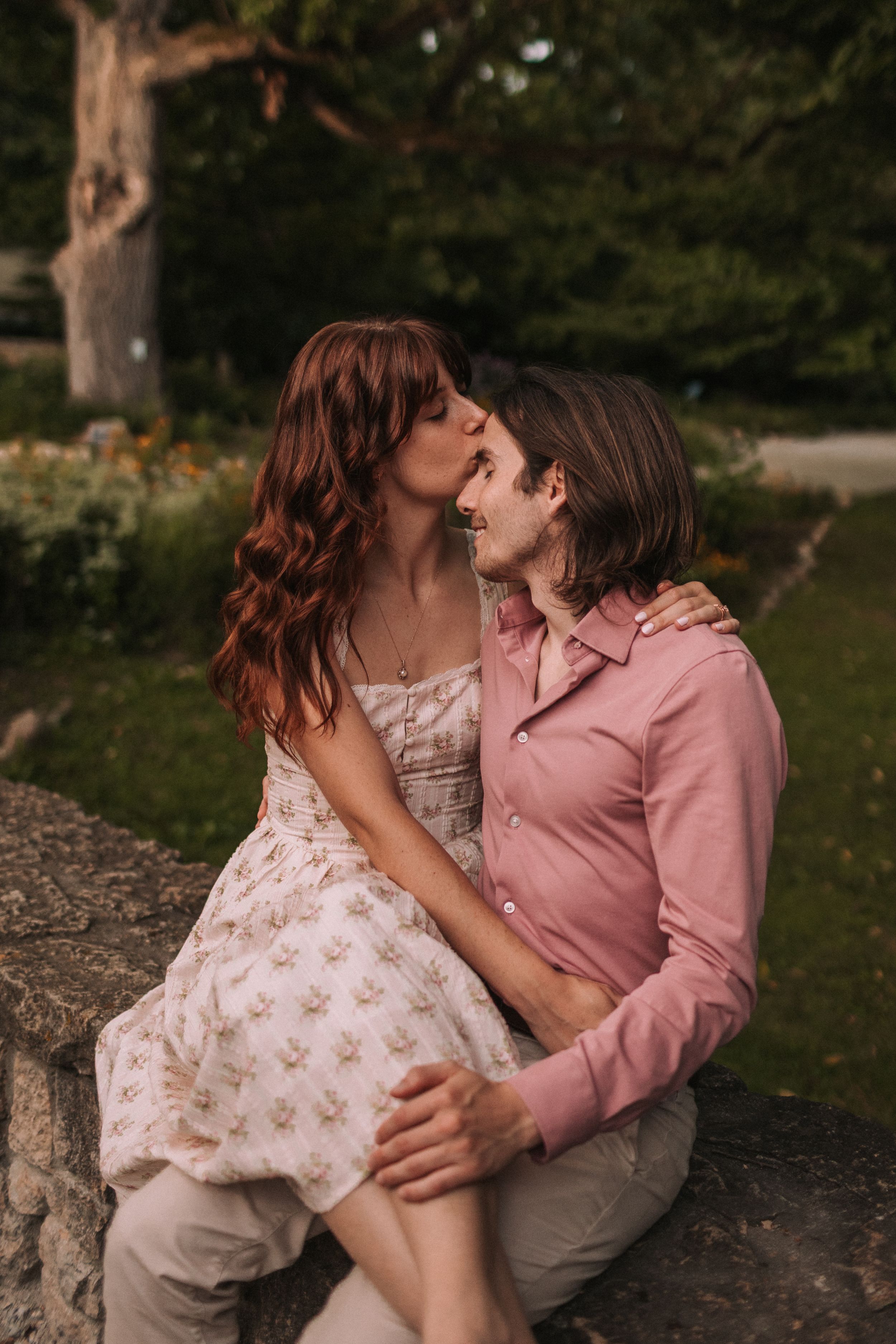 A romantic couple sits on a stone wall in a garden sharing intimate moments during a sunset photoshoot.
