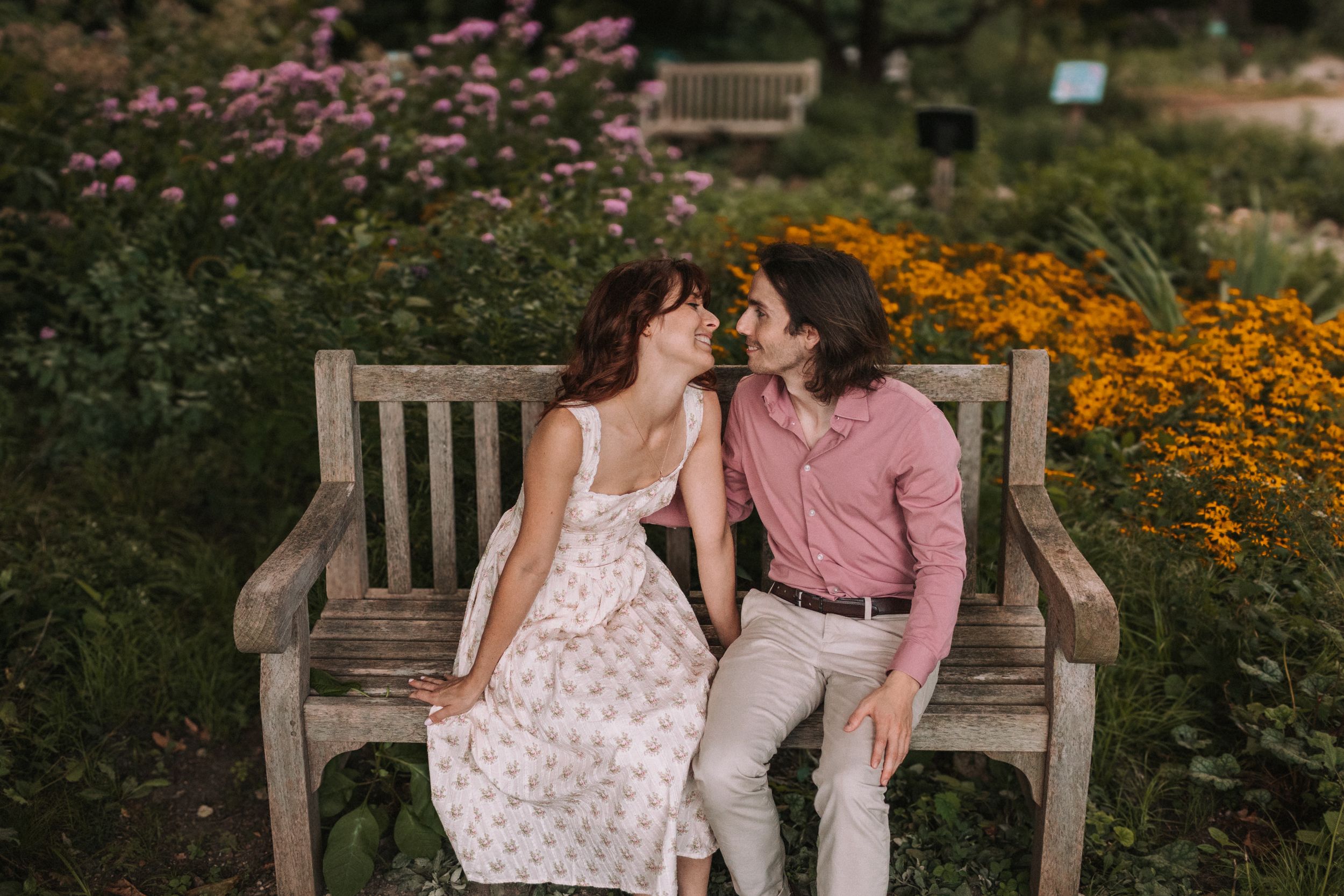 A couple sits on a wooden bench surrounded by vibrant orange and pink flowers in a lush garden setting.