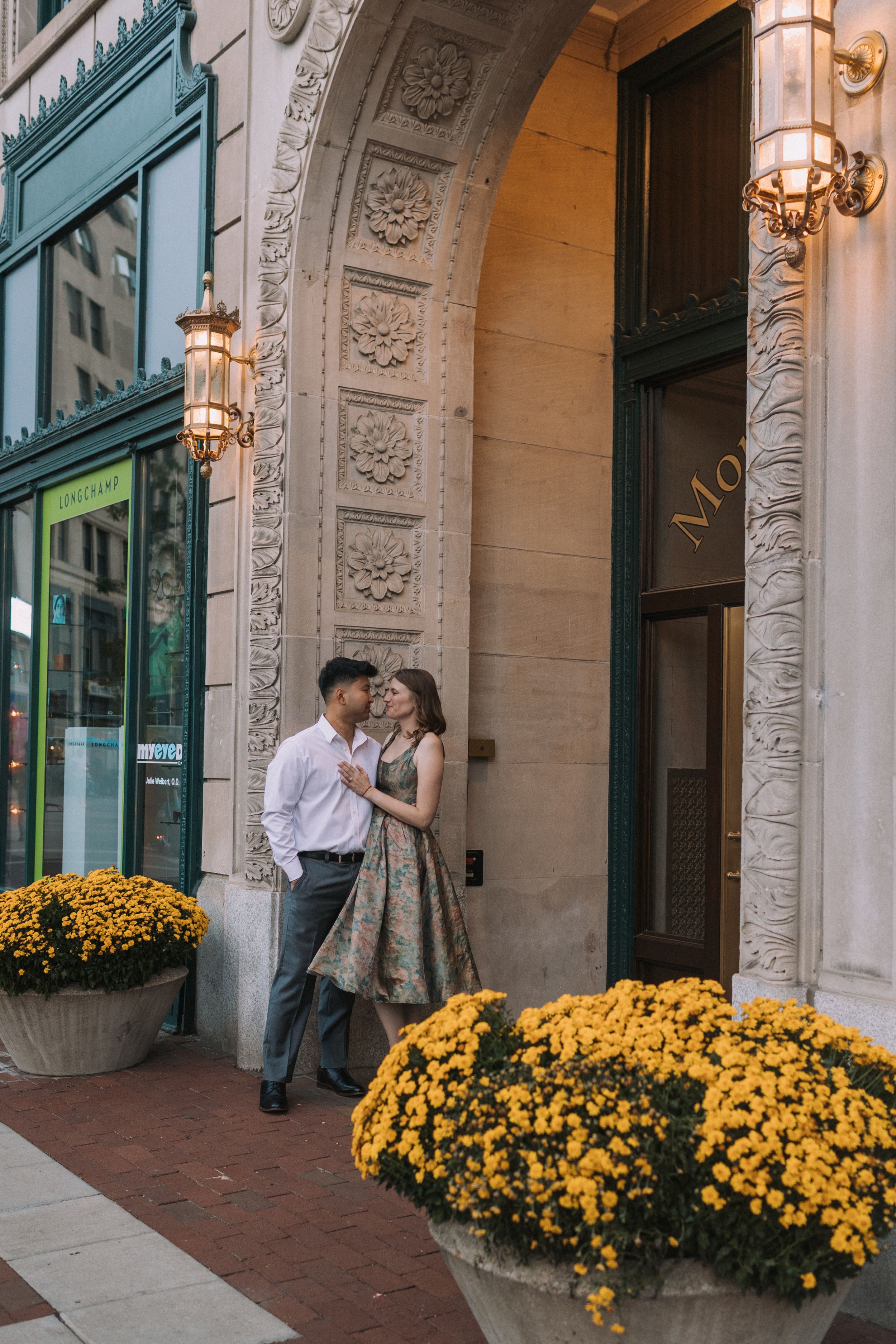 Romantic couple posing by yellow mums and classical architecture with warm evening lighting.