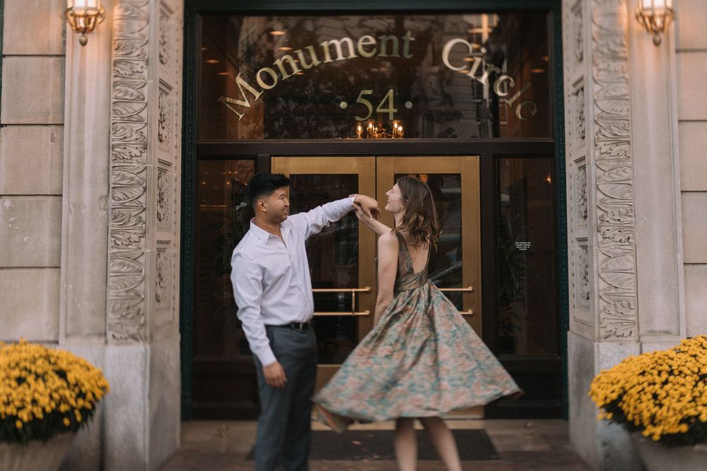 A couple in formal attire stands in front of a building entrance with yellow flowers and ornate decor.