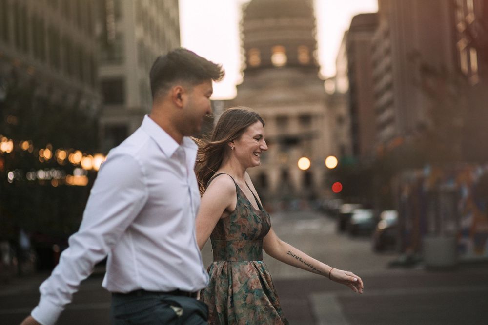 A couple embraces and dances together on an urban street at dusk with warm glowing lights in the background.