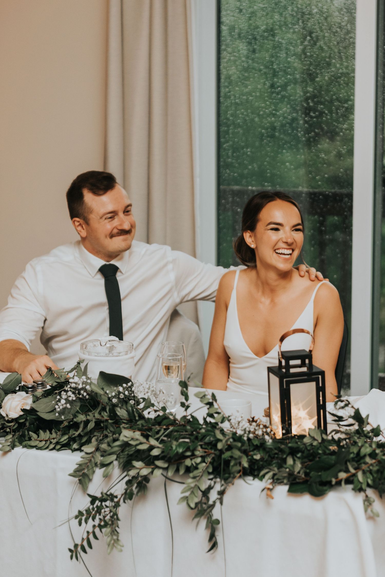 Wedding party members in white attire laugh together at reception table decorated with eucalyptus garland.