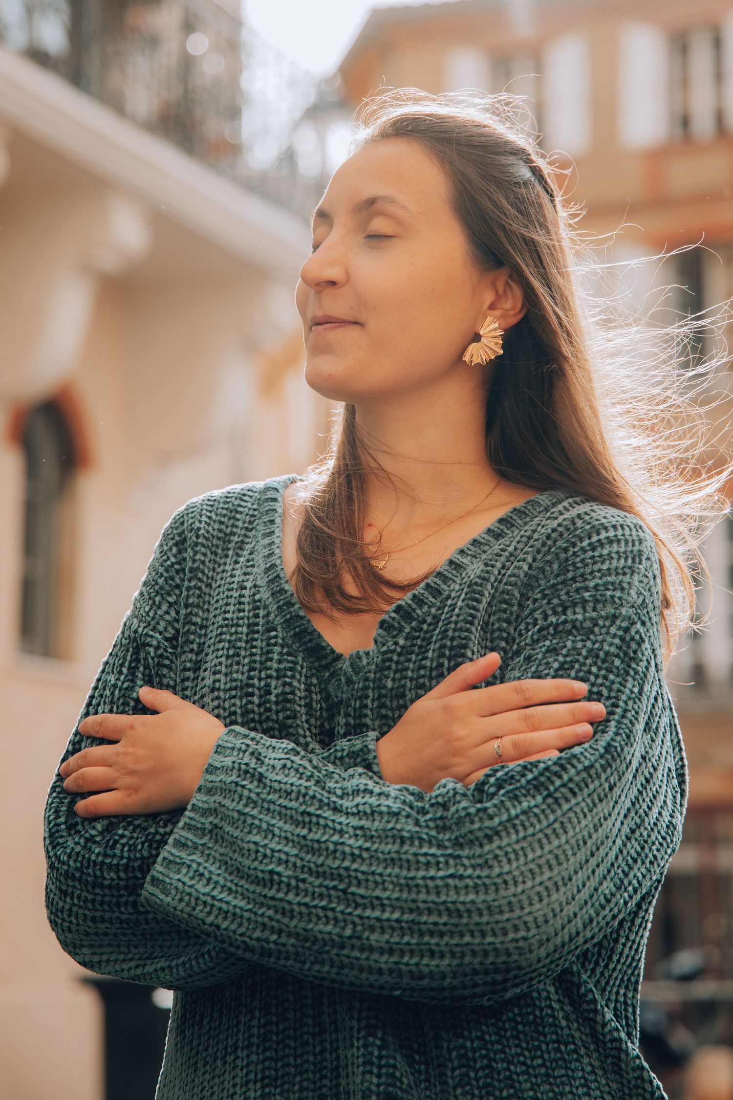 Woman in teal knit sweater standing with arms crossed and eyes closed in sunlight outdoors.