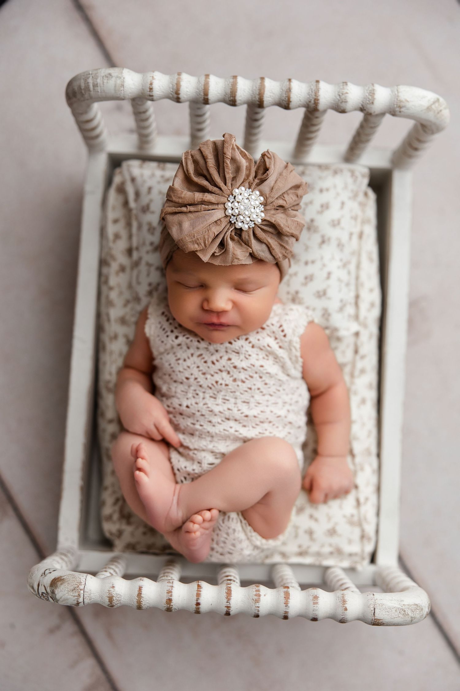 Newborn baby wearing white lace romper and tan floral headband sleeps peacefully in vintage white wooden bed prop.