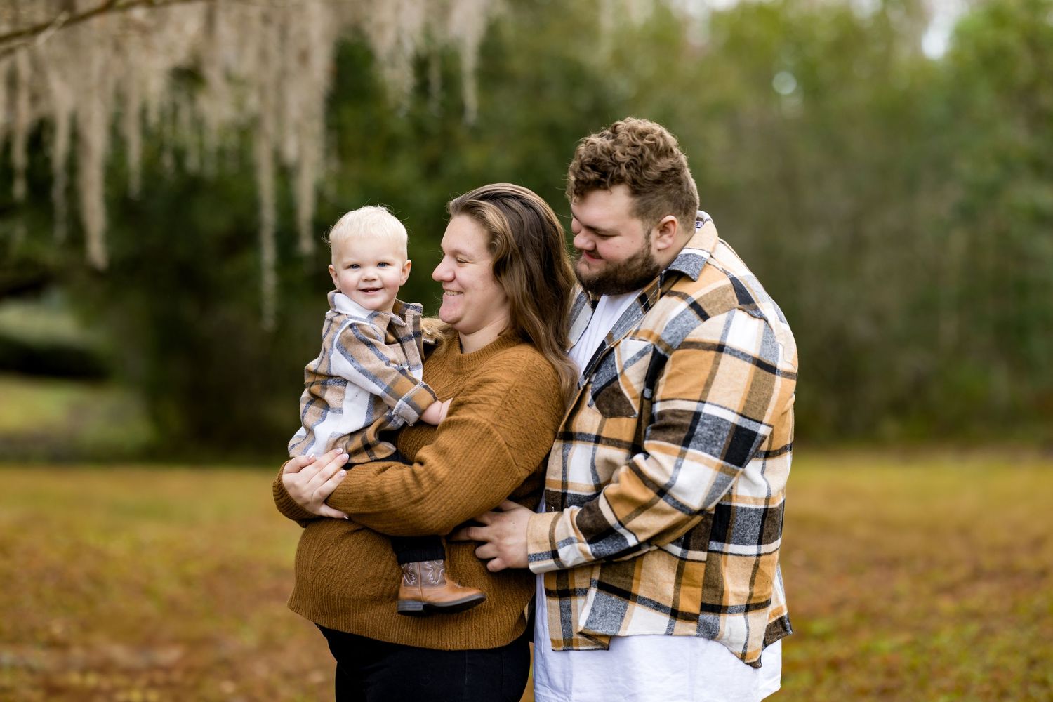 Family wearing coordinating brown and plaid outfits embraces while laughing together in an autumn field setting.