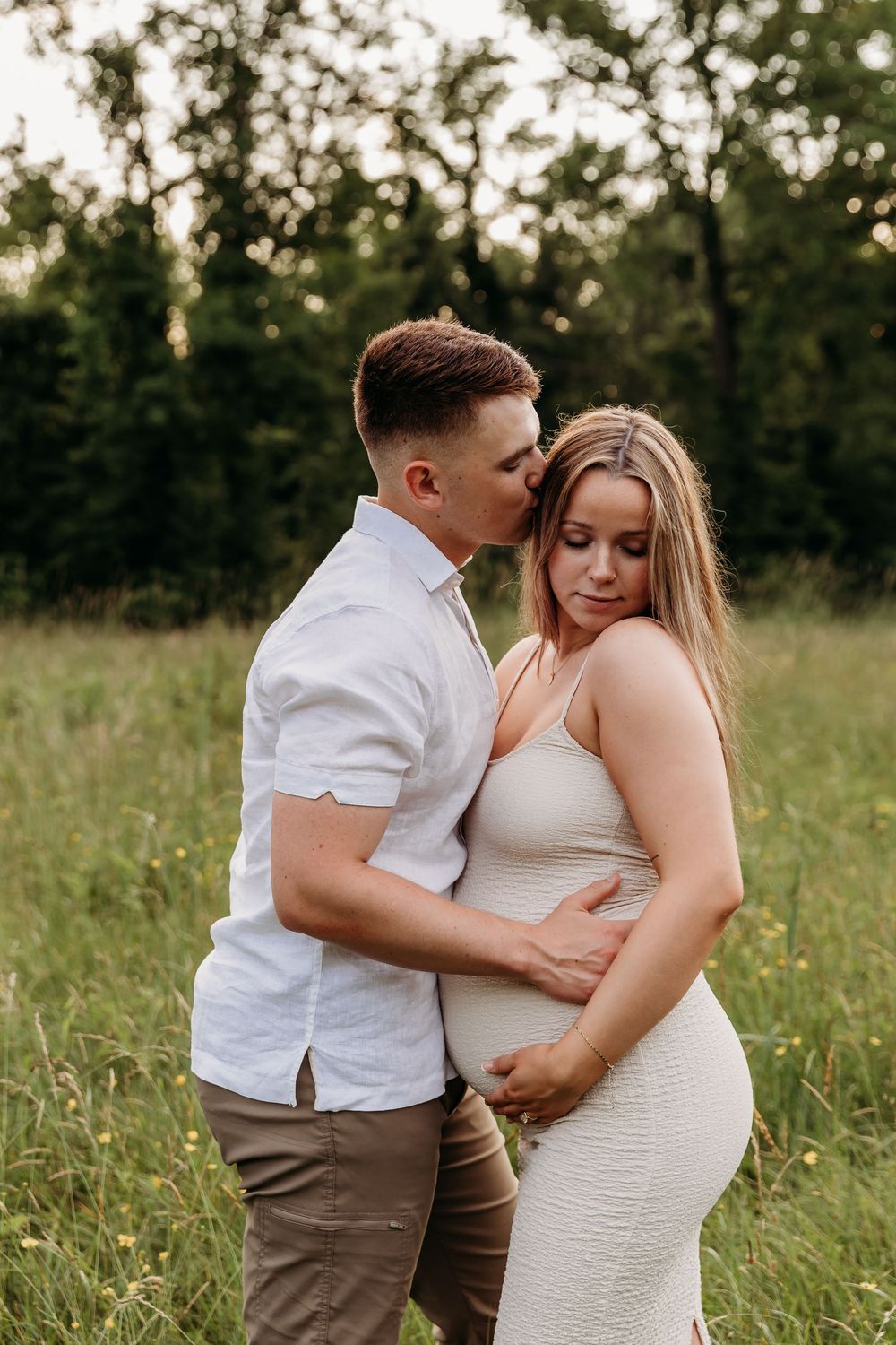 A couple in white and brown clothing embraces in a field during a maternity photoshoot at sunset.