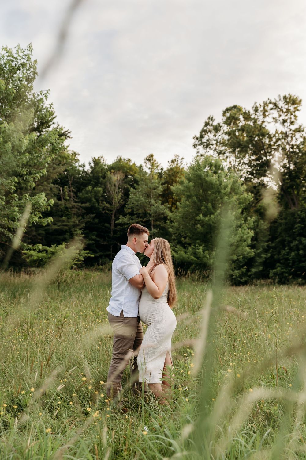 A romantic couple embraces in a sunlit meadow with tall grass and trees in the background.