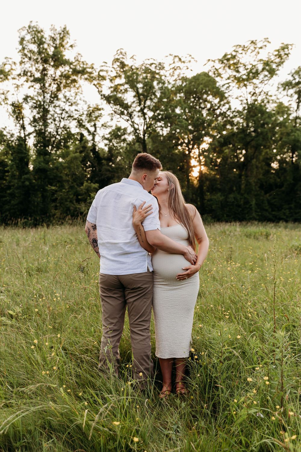 A couple shares an intimate moment in a golden meadow at sunset while wearing white and brown casual attire.