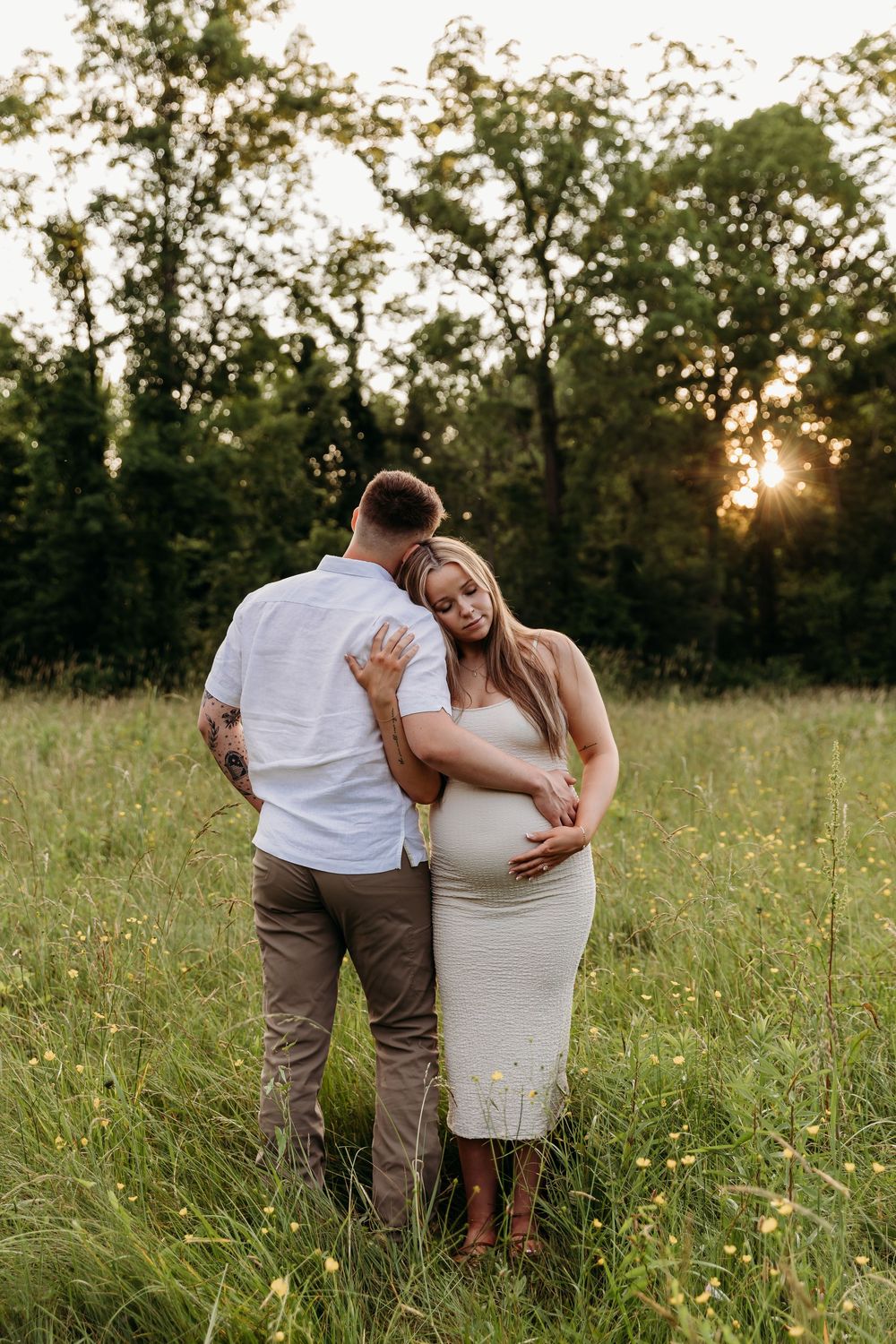 A couple shares an intimate moment in a sunlit meadow during a maternity photo session at golden hour.