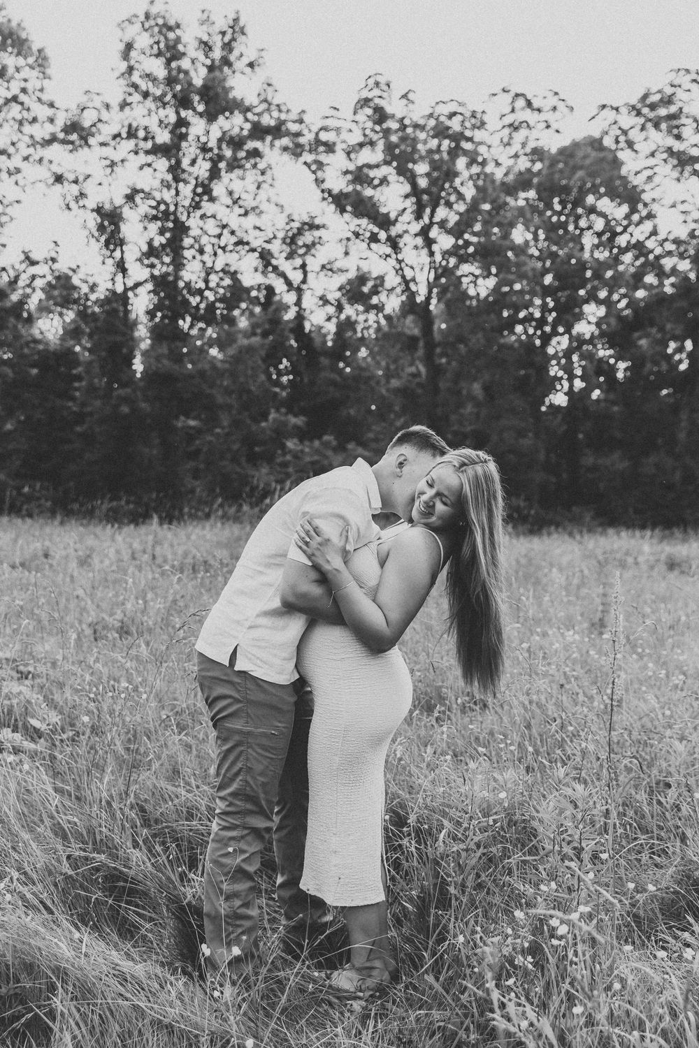 A romantic couple shares an intimate embrace in a sunlit meadow with tall grass and trees in the background.