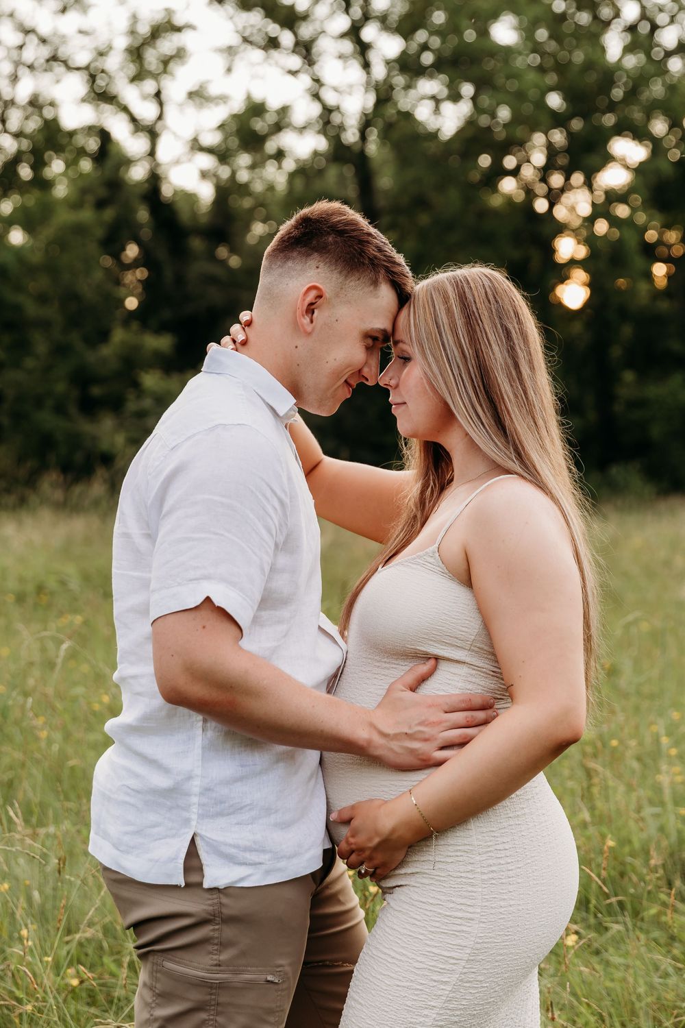 A couple embraces tenderly in a sunlit meadow wearing white and tan clothing during a romantic photoshoot.