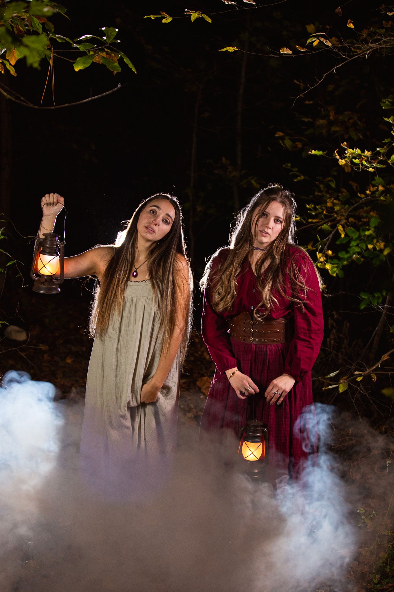 Two women pose with lanterns in a dark forest setting as smoke creates an eerie atmosphere.