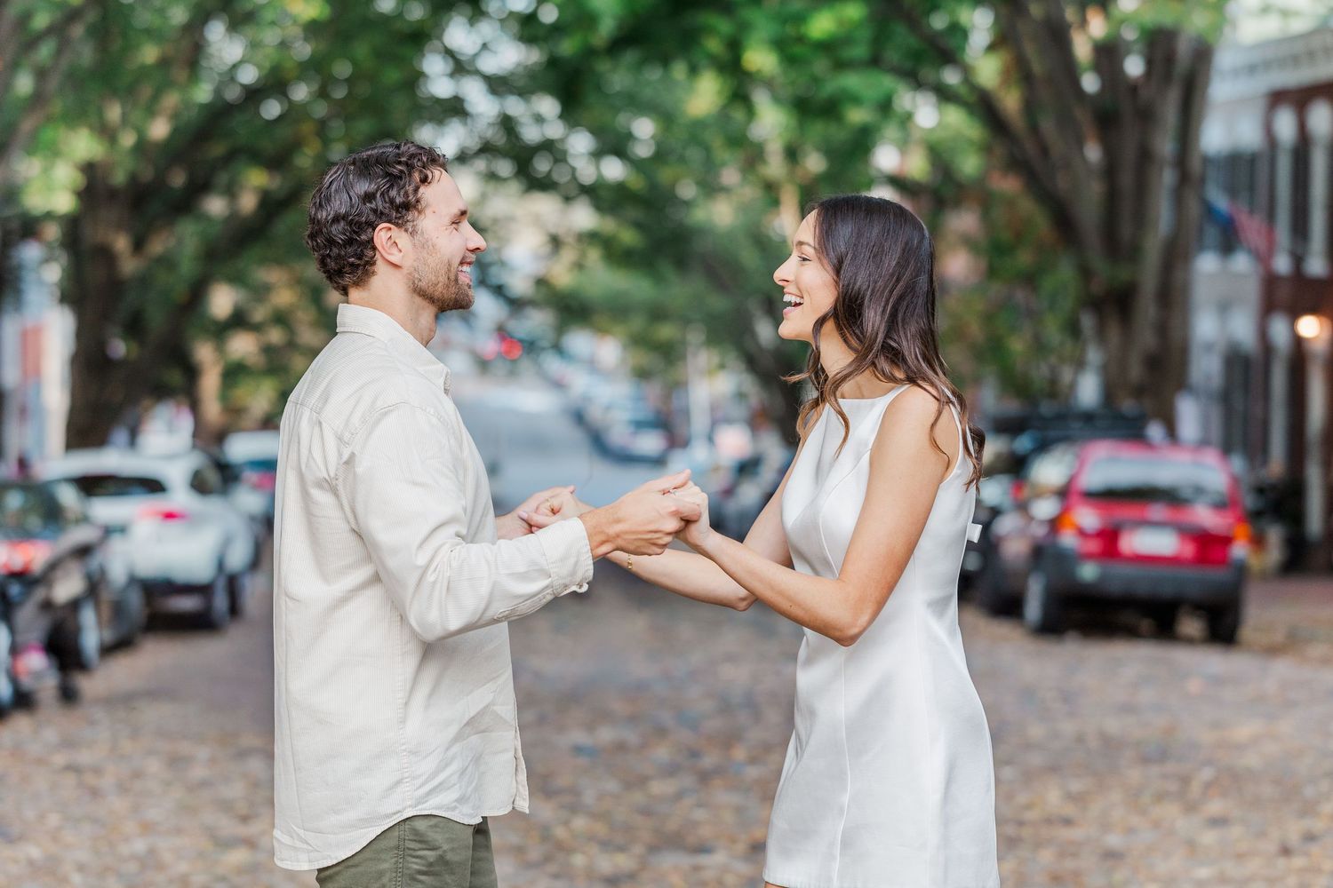A couple in white outfits shares a romantic moment dancing together on a tree-lined street.
