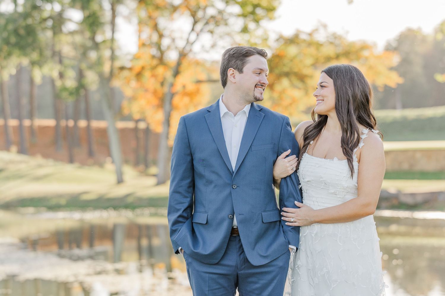 A couple in wedding attire shares a romantic moment by a pond surrounded by autumn trees at sunset.