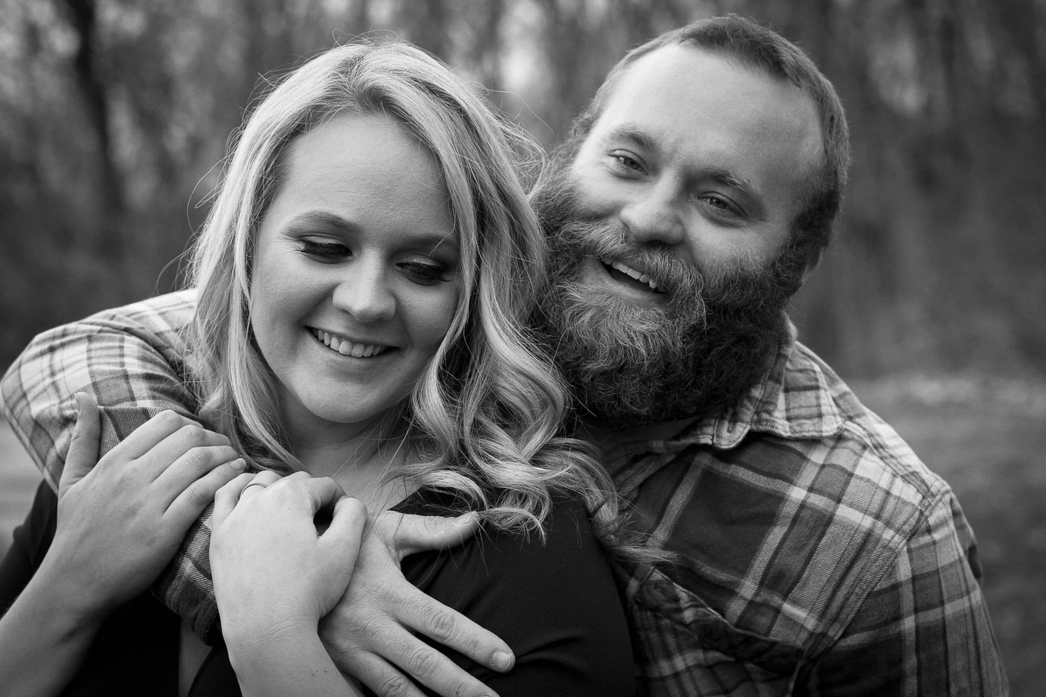 A woman smiles, looking down as her fiance grins at the camera in a black-and-white edit from a KC engagement photo session.