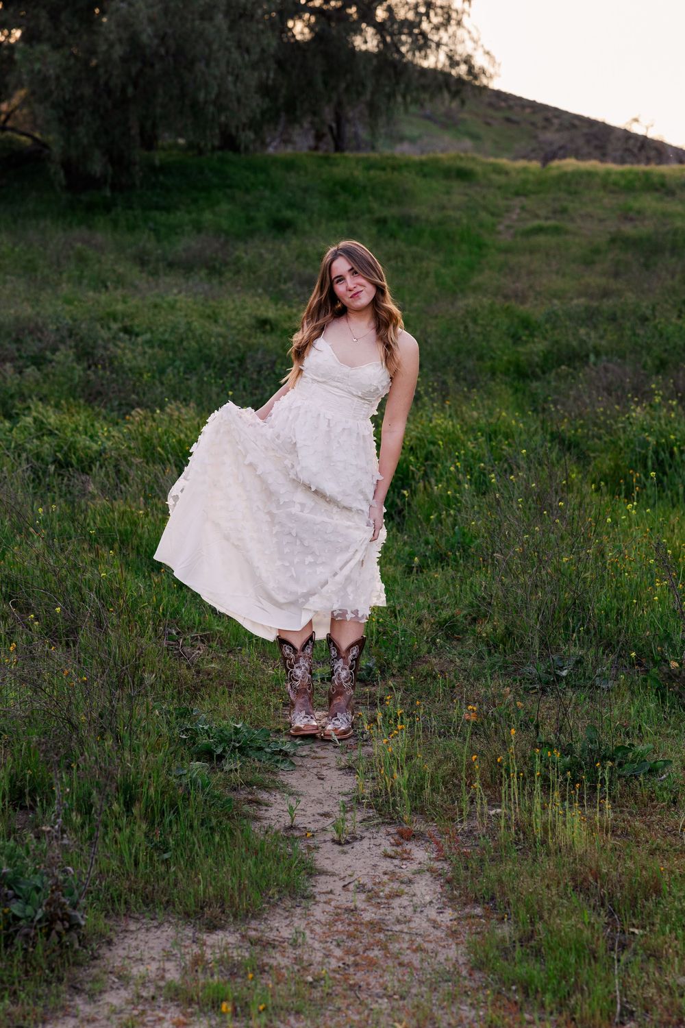 A person in a flowing white lace dress and boots poses on a grassy hillside path at sunset.