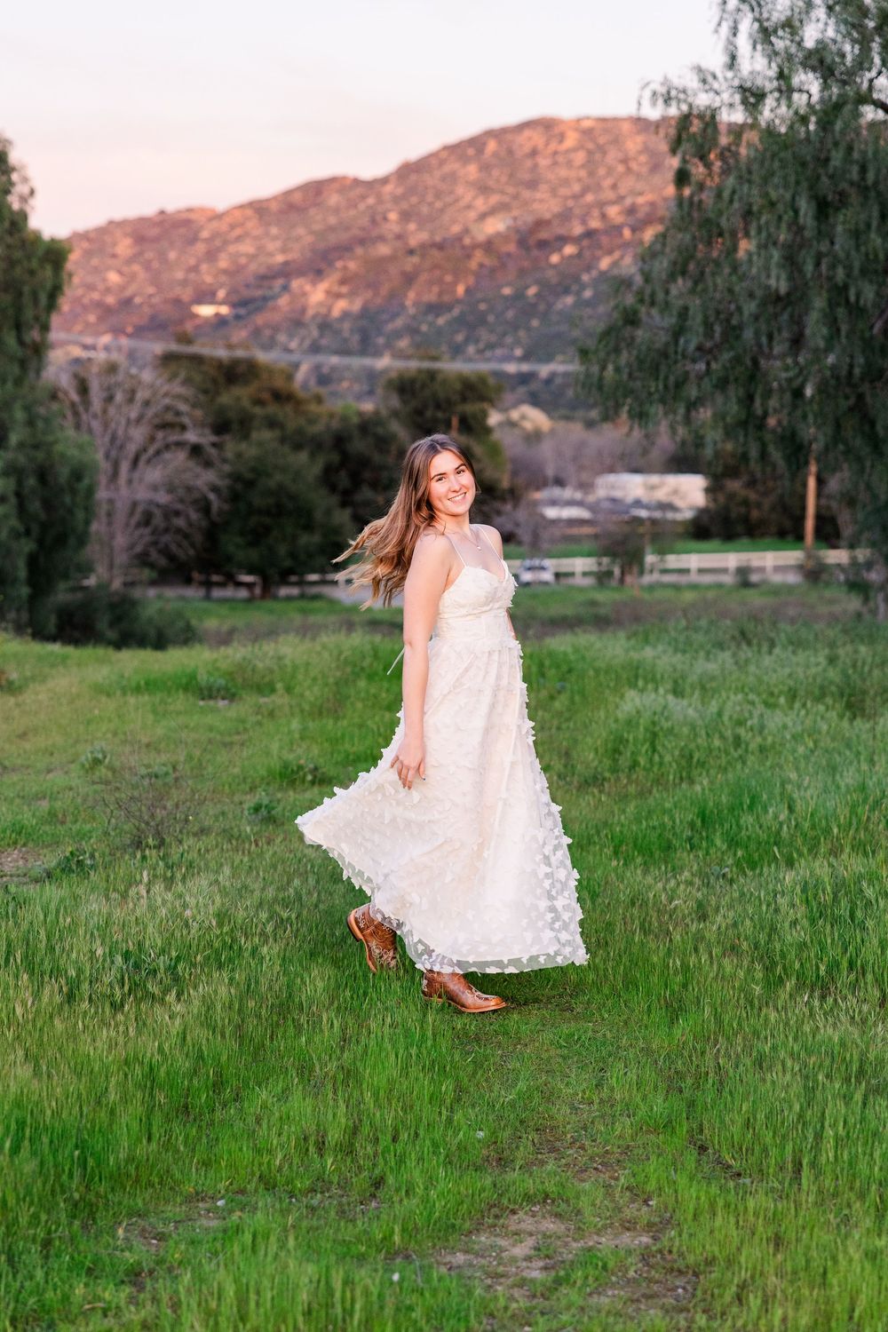 A child in a white lace dress twirls playfully in a green meadow with mountains visible in the sunset background.
